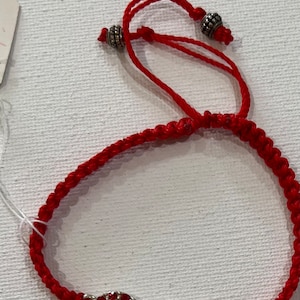 May include: Red string bracelet with an infinity symbol charm. The charm is made of silver and has red crystals.