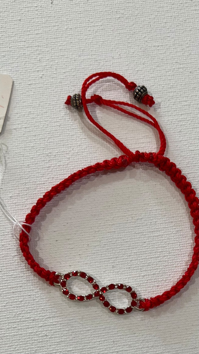 May include: Red string bracelet with an infinity symbol charm. The charm is made of silver and has red crystals.