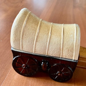 Puede incluir: Una figurita de cerámica de una carreta cubierta con ruedas marrones y una lona beige. La carreta tiene una caja de madera en la parte trasera.