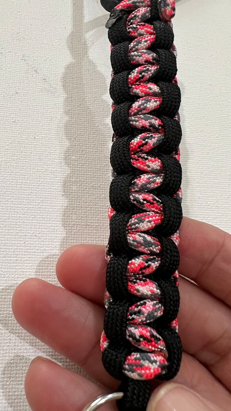 K&ouml;nnte beinhalten: Ein schwarzer und pinkfarbener Paracord-Schl&uuml;sselanh&auml;nger mit einem silbernen Schl&uuml;sselring. Der Paracord ist in einem komplexen Muster gewebt.