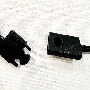 K&ouml;nnte beinhalten: Zwei schwarze Plastikverschl&uuml;sse f&uuml;r eine Schnur oder ein Band. Ein Verschluss hat ein kleines Loch in der Mitte.