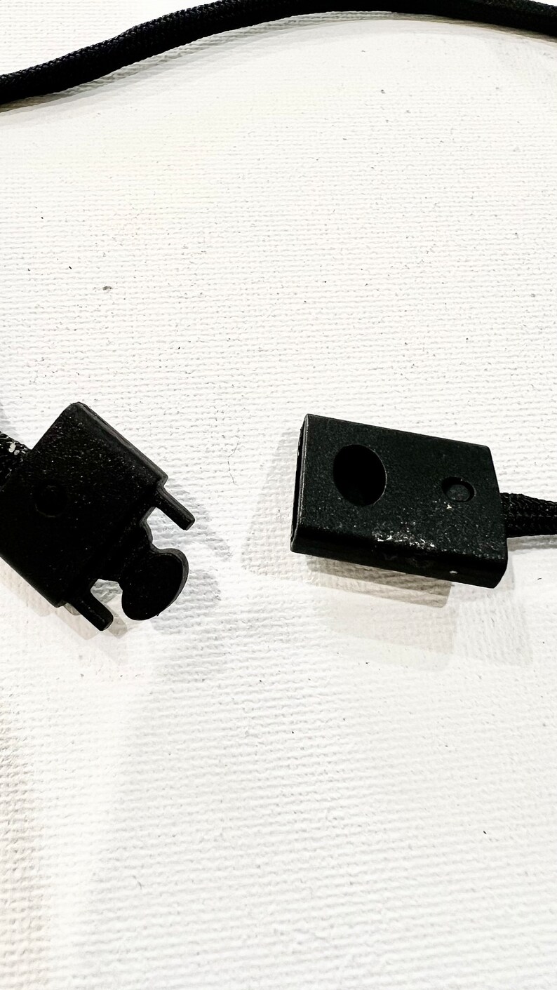 K&ouml;nnte beinhalten: Zwei schwarze Plastikverschl&uuml;sse f&uuml;r eine Schnur oder ein Band. Ein Verschluss hat ein kleines Loch in der Mitte.