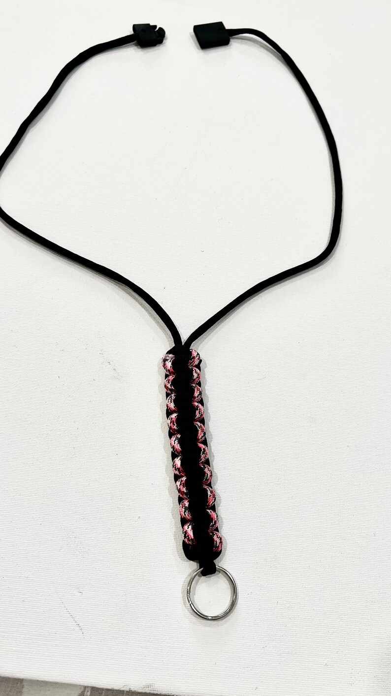 K&ouml;nnte beinhalten: Ein schwarzes Lanyard mit einem pinkfarbenen und schwarzen Paracord-Knoten und einem silbernen Schl&uuml;sselring.
