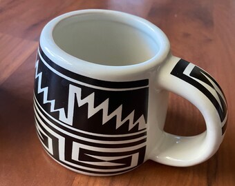 Puede incluir: Taza de cerámica blanca con un patrón geométrico en blanco y negro. El patrón presenta zigzags y cuadrados.