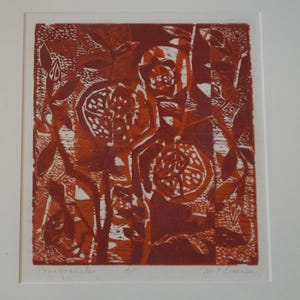 May include: A framed block print featuring a stylized depiction of pomegranates and foliage in shades of deep red and burnt orange. The artwork is set within a white mat and a dark brown frame, with handwritten text visible at the bottom edge.