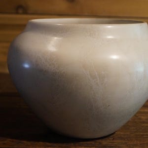 May include: A cream-colored, rounded ceramic vase with a wide opening. The vase has a smooth, glossy finish with subtle, textured veining across its surface. The vase sits on a wooden surface, with a wooden background.