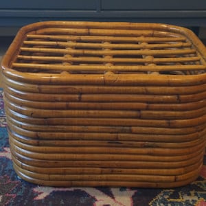 May include: A square, low-profile rattan coffee table. The table is constructed from curved, horizontal bamboo strips, with a slatted top. The natural wood tones and rounded edges give it a warm, inviting aesthetic. The table measures approximately 24 inches wide and 18 inches tall.