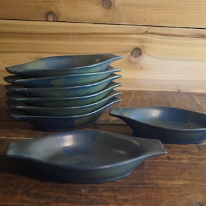 May include: A stack of eight blue-green ceramic serving dishes with handles, alongside two additional dishes. The dishes have a unique, elongated shape and are arranged on a wooden surface, with a wooden background.