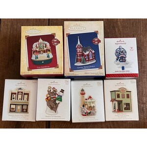 May include: A collection of nine Hallmark Keepsake ornaments, each featuring a different holiday scene. The ornaments are packaged in individual boxes with a white background and red and blue accents. The ornaments include a city scene, a church, a Ferris wheel, a police station, a tree with ornaments, a lighthouse, and a store.