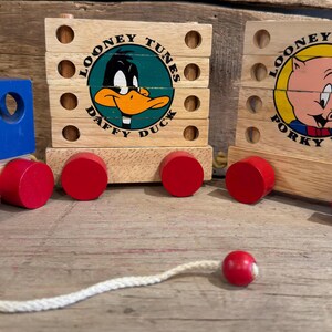 May include: Wooden Looney Tunes train toy featuring Daffy Duck and Porky Pig. The train has red, blue, and yellow accents, with a white pull string and red wheels. The toy is made of wood and is designed for children.