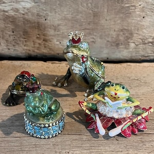 May include: Four frog figurines, each with a different pose and detail. One frog wears a crown and holds a jewel, another is a ballerina with a red and white tutu, and two others are sitting with jeweled accents.