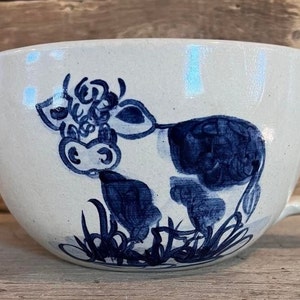 May include: A white ceramic mixing bowl with a blue and white cow design. The bowl has a handle and a spout.