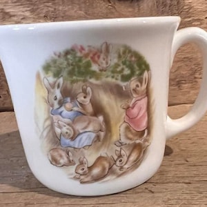 May include: White ceramic mug with a handle. The mug features a colorful illustration of Peter Rabbit and his friends from Beatrix Potter's children's book.