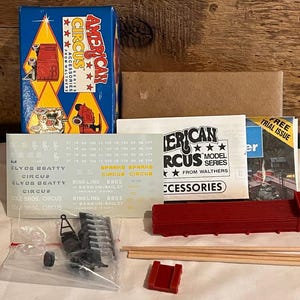 May include: A model circus train set with various components, including a red flatbed car, wooden dowels, and a blue box with "American Circus" text. Also included are decals and a small instruction booklet.