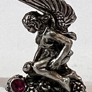 May include: A silver-toned figurine of a winged figure kneeling on a textured base. The figure has detailed wings and is posed with a bent posture. A round, red gemstone is set into the base.