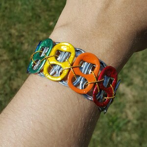 May include: A rainbow colored bracelet made from recycled aluminum pull tabs. The bracelet is made of six pull tabs, each painted a different color of the rainbow. The pull tabs are connected together with wire.