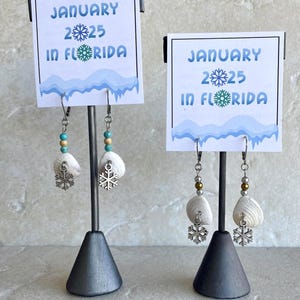 May include: Earrings featuring white seashells, turquoise and gold beads, and silver snowflake charms. The display stand has a sign that reads "JANUARY 25 IN FLORIDA".
