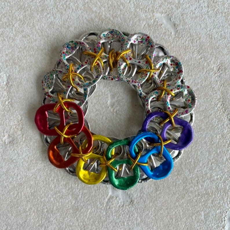 May include: A rainbow-colored wreath made from pull tabs. The tabs are connected together with wire and have a sparkly finish.
