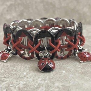 Könnte beinhalten: Ein silberfarbenes Armband mit einem sich wiederholenden Muster aus schwarzen und roten Akzenten. Das Armband ist mit roten Marienkäfer-Anhängern verziert. Die Marienkäfer haben rote Körper mit schwarzen Punkten und schwarzen Köpfen. Das Armband ist mit roter Kordel verbunden.