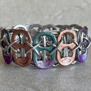 Puede incluir: Un brazalete hecho a mano con lengüetas de latas de aluminio recicladas. El brazalete presenta una mezcla de colores metálicos, incluyendo cobre, verde azulado y púrpura, con un acabado brillante en algunas lengüetas. Las lengüetas están unidas con alambre plateado.