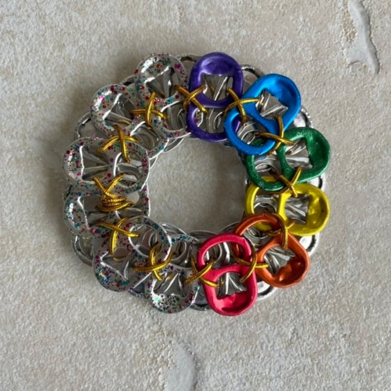 May include: A colorful wreath made from pull tabs from aluminum cans. The tabs are arranged in a circle and are connected with wire. The tabs are painted in the colors of the rainbow.