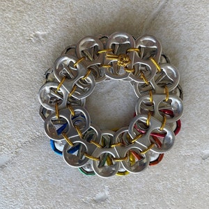 May include: A circular wreath made from aluminum pull tabs, connected with yellow string. The pull tabs are arranged in a circle, with the tops facing outwards. The wreath is a colorful and unique craft project.