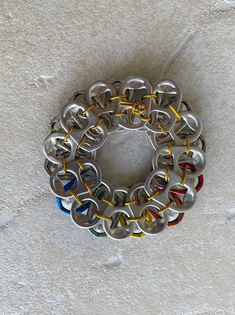 May include: A circular wreath made from aluminum pull tabs, connected with yellow string. The pull tabs are arranged in a circle, with the tops facing outwards. The wreath is a colorful and unique craft project.