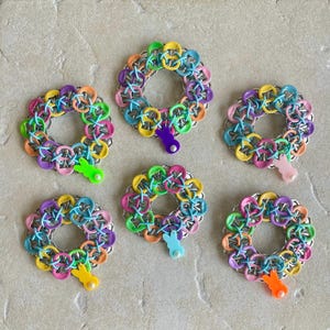 May include: Six colorful wreaths made of linked metal rings, each with a pastel-colored bunny charm hanging from the bottom. The wreaths are in shades of pink, blue, green, yellow, and purple.