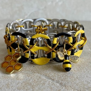 May include: A yellow and black bee-themed bracelet made from recycled aluminum can tabs. The bracelet has a honeycomb charm and a bee charm with rhinestones.