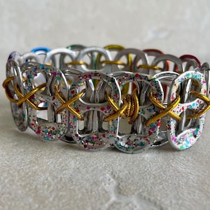 May include: A silver bracelet made from pull tabs with gold wire woven through the loops. The pull tabs are covered in colorful glitter.