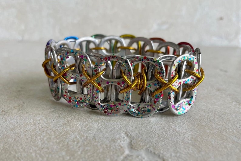 May include: A silver bracelet made from pull tabs with gold wire woven through the loops. The pull tabs are covered in colorful glitter.