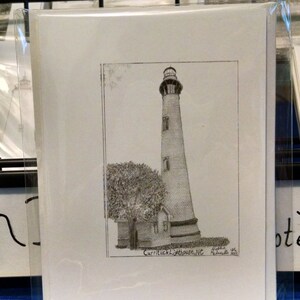 Op de afbeelding: Een zwart-wit potloodtekening van de Currituck Lighthouse in North Carolina, met een boom en een klein gebouw. De tekening staat op wit papier en is gesigneerd. Het kunstwerk zit in een transparante plastic hoes.