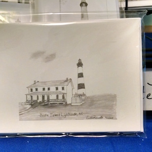 Op de afbeelding: Een potloodschets van de Bodie Island Lighthouse in North Carolina, met de vuurtoren met zwart-witte strepen en een aangrenzend gebouw. Het kunstwerk staat op een witte kaart, verpakt in een transparante plastic hoes.
