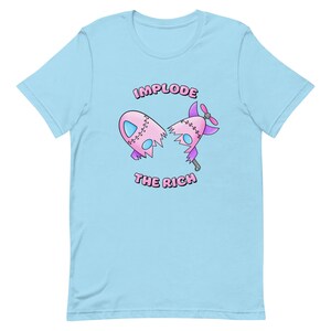 May include: A light blue t-shirt with a pink and purple cartoon rocket ship that is broken in half. The text "IMPLODE THE RICH" is printed on the shirt.
