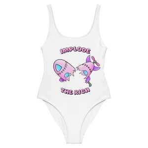 May include: White one-piece swimsuit with a pink and purple cartoon rocket ship that is broken in half. The text "Implode the Rich" is printed on the swimsuit.