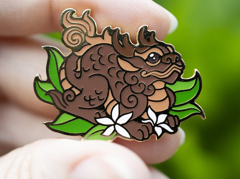 Puede incluir: Un pin de esmalte marr&oacute;n y dorado que representa una criatura m&iacute;tica con una cola enroscada, cuernos y un dise&ntilde;o floral. La criatura est&aacute; rodeada de hojas verdes y flores blancas.