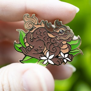Puede incluir: Un pin de esmalte marr&oacute;n y dorado que representa una criatura m&iacute;tica con una cola enroscada, hojas verdes y flores blancas. La criatura tiene un dise&ntilde;o detallado con remolinos y patrones.