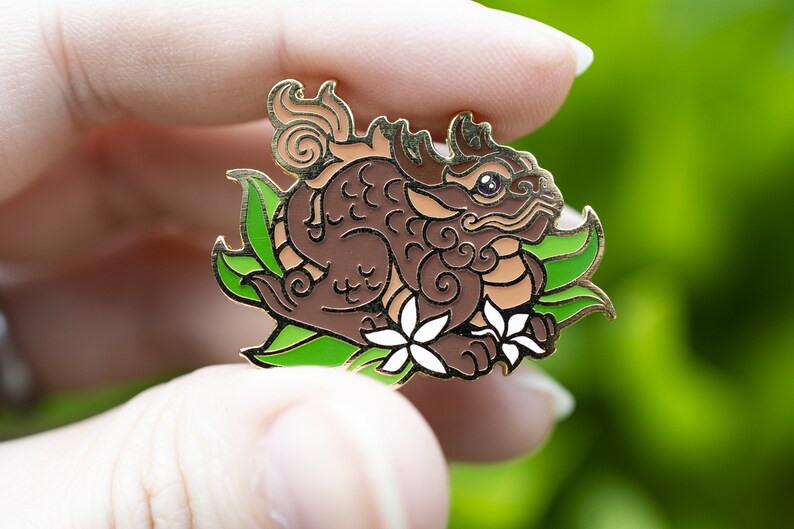 Puede incluir: Un pin de esmalte marr&oacute;n y dorado que representa una criatura m&iacute;tica con una cola enroscada, hojas verdes y flores blancas. La criatura tiene un dise&ntilde;o detallado con remolinos y patrones.