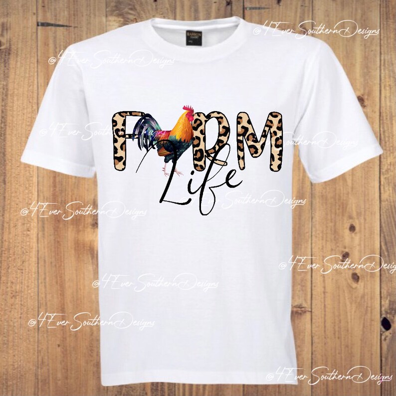 May include: White t-shirt featuring the text "FARM LIFE" with a colorful rooster graphic. The word "FARM" has a leopard print design. The shirt is a crew neck and has short sleeves.