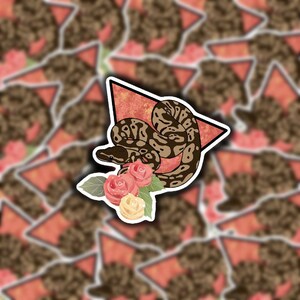 May include: A sticker featuring a brown and tan snake coiled around a bouquet of pink and yellow roses. The snake is set against a red triangle with a white border. The background is a pattern of similar stickers.