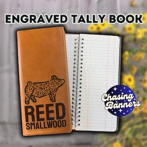 May include: An open, tan leather tally book with a spiral binding. The cover features a black engraved illustration of a pig with a spotted pattern, the name "REED SMALLWOOD", and the text "ENGRAVED TALLY BOOK". The notebook is open to lined pages.