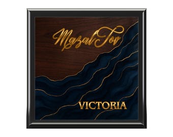 May include: Square black framed art piece with a dark wood grain background and a navy blue and gold wave design. The words "Mazal Tov" and "Victoria" are written in gold script.