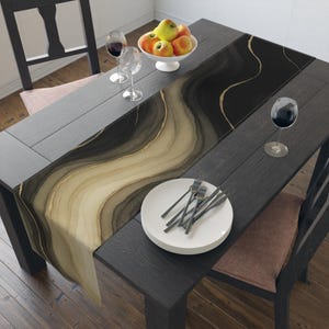 May include: A dark wooden dining table is set with a decorative table runner featuring a flowing, abstract design in shades of black, brown, and gold. The table is set with plates, silverware, wine glasses, and a bowl of apples.