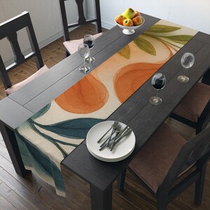 May include: A dining table set for a meal, featuring a watercolor-style table runner with orange and teal leaf designs. The table is set with plates, silverware, wine glasses, and a bowl of fruit. The table and chairs are dark wood.