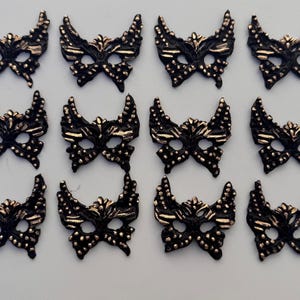 May include: Twelve black and gold butterfly-shaped masks. Each mask has a detailed design with gold accents and small, raised dots. The masks are arranged on a white surface, suitable for crafting or costume use.