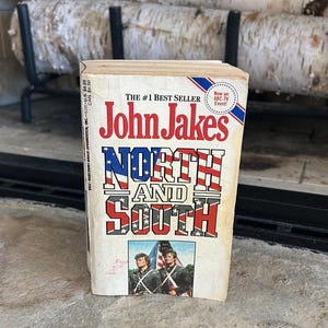 May include: A paperback book titled "North and South" by John Jakes. The cover features the title in red, white, and blue, with an image of two men in military uniforms. The book is a #1 bestseller and was an ABC-TV event.