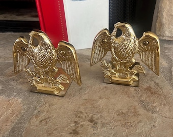 May include: A pair of polished brass bookends, each featuring a detailed eagle with wings spread wide, perched atop a base. The eagles are depicted in a classic heraldic style with textured feathers and are positioned symmetrically on a stone surface.