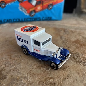 May include: A white and blue toy truck featuring the Houston Astros logo on top and the text "Houston Astros" on the side. The year "1990" is printed in red. The truck has a classic design with a blue front bumper.