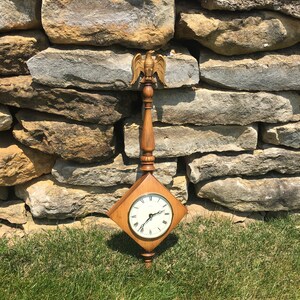 May include: A wooden clock with a square face and a gold eagle finial. The clock is hanging on a stone wall with green grass in the foreground.