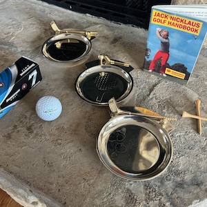 May include: Three silver-toned golf ashtrays with gold-colored golf club accents. A golf ball, a box of golf balls, a golf handbook, and golf tees are also present. The ashtrays are round with a black interior.